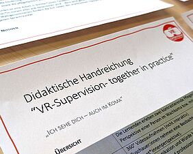 Eine Detailaufnahme zeigt einen Ausschnitt eines Printdokuments. Auf dem Papier steht „Didaktische Handreichung – VR-supervision – together in practise“.