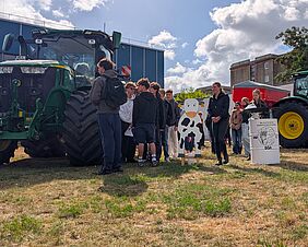 Eine Gruppe junger Menschen betrachtet einen Traktor. Sie stehen auf einer Wiese. Hinter ihnen befindet sich ein blockartiges Gebäude. Der Himmel ist strahlend-sonnig mit großen hellen Wolken.