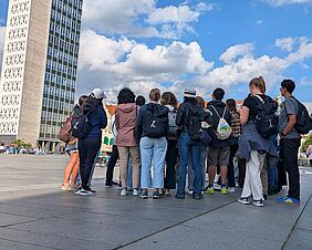 Eine Gruppe junger Menschen steht mit dem Rücken zur Kamera. Sie stehen auf dem Neubrandenburger Marktplatz und schauen auf das Haus der Kultur und Bildung. Der Himmel ist satt blau mit hellen weißen Wolken.