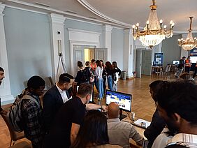 In einem hellen festlichen Saal tummeln sich Personen an verschiedenen Informationsständen. Im Vordergrund befindet sich eine Menschentraube, die gemeinsam auf einen Monitor schaut. Eine der Personen zeigt auf den Bildschirm.