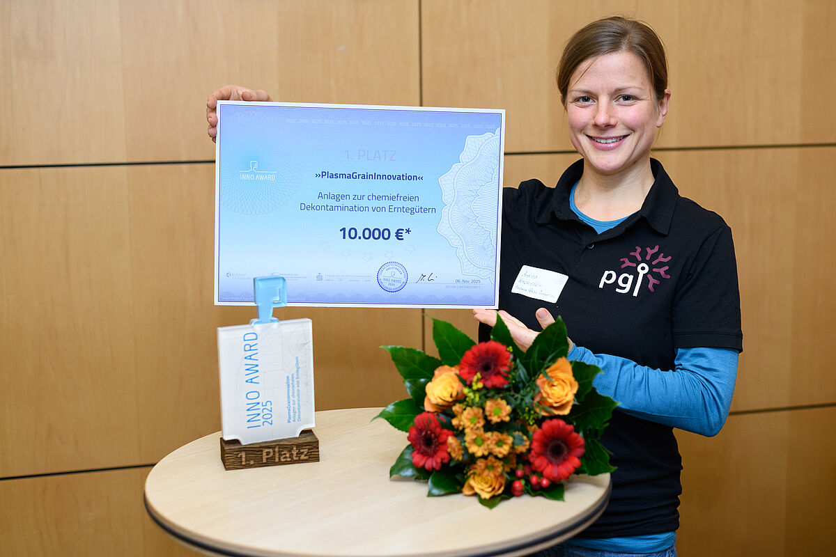 Eine Frau lächelt freundlich in die Kamera. Sie trägt ein schwarzes Poloshirt mit der Aufschrift „pgi“, darunter ein blaues Langarmshirt. In der Hand hält sie einen großen weißen Bilderrahmen, in dem sich eine Art symbolischer Check mit der Aufschrift „10.000 Euro“ befindet. Vor ihr befindet sich ein runder Stehtisch mit einem rot-gelben Blumenstrauß. Außerdem befindet sich der sogenannte Inno Award auf dem Tisch – ein viereckiger, gläserner Preis auf einem hölzernen Sockel.