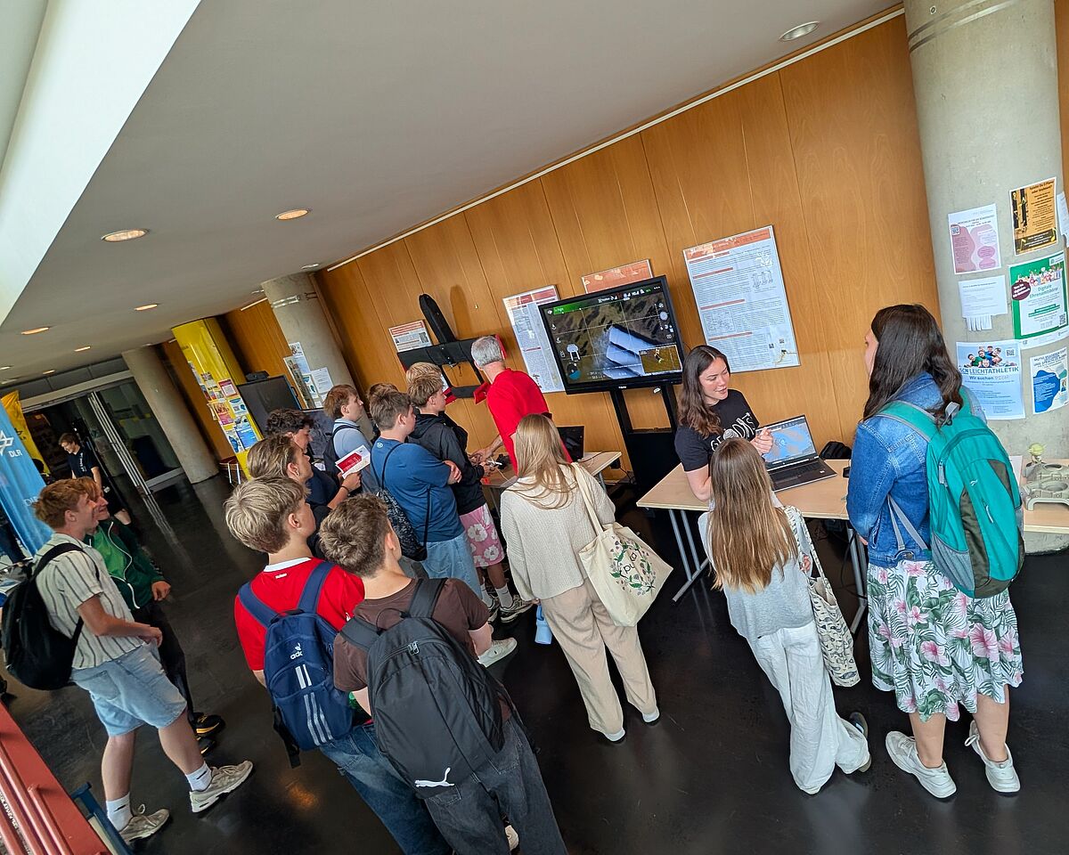 Es herrscht Trubel in einem Foyer. Die auf dem Foto dargestellte Situation erinnert an eine Messe. Schülerinnen und Schüler mit Rucksäcken und Taschen stehen an Informationsständen und unterhalten sich mit den Ausstellenden.