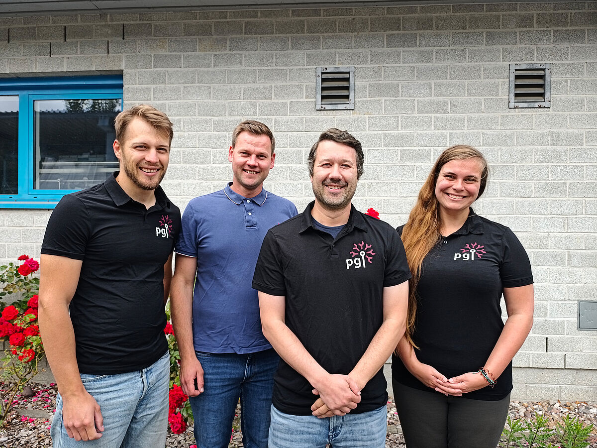 Vier Personen lächeln freundlich in die Kamera. Drei von ihnen tragen schwarze Shirts mit der Aufschrift „PGI“.