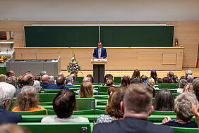 Ein Mann, der einen blauen Anzug und ein weißes Hemd trägt, steht vor einem Pult und einem voll besetzten Hörsaal. Hinter ihm befindet sich eine große grüne Kreidetafel. 