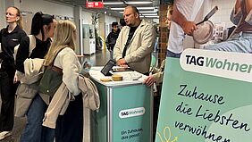 Zwei Frauen, vielleicht Mutter und Tochter, informieren sich an einem Messestand. Im Vordergrund befindet sich ein Rollup - ein Aufsteller, der über das Thema Wohnen informiert.