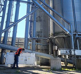 Ein Mann in orangefarbener Warnweste inspiziert eine Gerätschaft, die einer weißen Kommode ähnelt. An ihr hängt ein großer weißer Schlauch. Im Hintergrund befindet sich ein großes Silo sowie ein verzweigtes Metallrohrsystem.