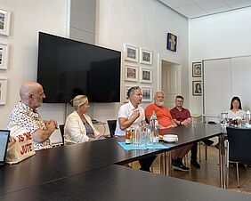 In einem historischen Saal mit holzvertäfelten Wänden, einem Kronleuchter und einem kunst-voll geschnitzten Balkon sitzen etwa 20 Menschen in mehreren Reihen auf hell gepolsterten Bänken. Alle blicken zur Kamera und lächeln. 