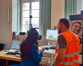 Eine Frau steht vor einem Monitor. Sie trägt eine VR-Brille. Neben ihr steht ein Mann, der eine orangefarbige Warnweste trägt.