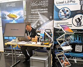 Das Foto zeigt einen Messestand des Unternehmens „Laser Scanning Europe“. Ein Mann sitzt an einem Laptop. Vor ihm ist ein Monitor zu sehen, der das Wort „Modellierung“ aufzeigt.