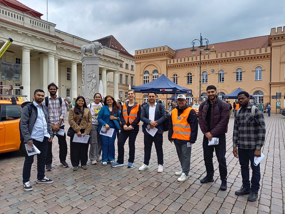 Zehn Personen stehen auf einem gepflasterten Marktplatz. Im Hintergrund ist das Rathaus von Schwerin zu erkennen. Die Personen sind locker gekleidet und lächeln in die Kamera. Zwei von ihnen tragen auffallende orangefarbige Warnwesten.