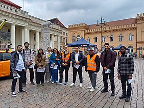 Zehn Personen stehen auf einem gepflasterten Marktplatz. Im Hintergrund ist das Rathaus von Schwerin zu erkennen. Die Personen sind locker gekleidet und lächeln in die Kamera. Zwei von ihnen tragen auffallende orangefarbige Warnwesten.