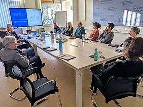 An einem Tisch in einem Seminarraum sitzen mehrere Personen an einem Tisch. Die Personen schauen gespannt auf ein blau-weißes Display mit Informationen zu digitalen Bildungsmaterialien..