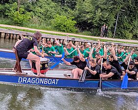 Zu sehen sind zwei Drachenboot-Teams in Aktion, Kopf an Kopf. Im Vordergrund befindet sich ein Team in Schwarz gekleidet, im Hintergrund tragen die Personen im Boot grüne Shirts. Auf dem Boot im Vordergrund sitzt eine Trommlerin, die ihrem Team den Takt angibt.