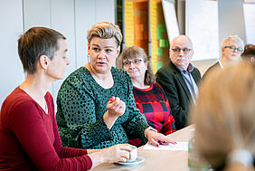 Eine blonde Frau mit großen Ohrringen sitzt wahrscheinlich an einem Konferenztisch. Sie trägt ein grünes Kleid mit schwarzen Punkten, hält einen Kugelschreiber in der Hand und spricht zu einer anderen Person. Hinter und vor ihr sind weitere Personen unscharf im Vorder- und Hintergrund zu erkennen. An den Wänden befinden sich verschiedenfarbige Heftnotizen. Weiße Flipcharts stehen im Hintergrund.