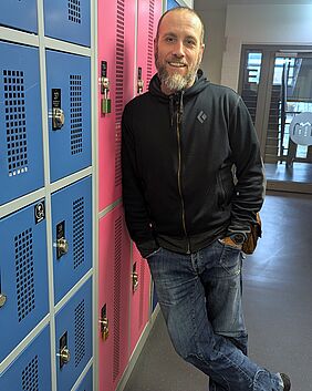 Der interviewte Reimar Goldschmidt lehnt lässig an einer Reihe bunter Spinte. Er lächelt freundlich und trägt lockere Kleidung. Die Spintfächer sind blau und rosa.