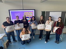 Acht Frauen stehen nebeneinander und lächeln in die Kamera. Hinter ihnen befindet sich ein Monitor, der die Aufschrift „Mentoring-Programm“ zeigt und eine Tafel, auf der „Herzlich willkommen“ zu lesen ist. Die Frauen tragen große einheitliche Taschen.