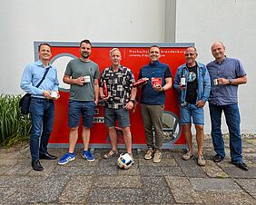 Sechs Männer posieren locker vor einer roten Torwand. Sie halten Tassen, Trinkflaschen und einen kleinen Pokal in ihren Händen. Vor ihnen liegt ein weiß-blauer Fußball. Alle lächeln freundlich in die Kamera.