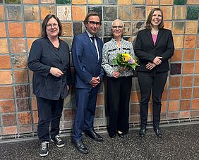 Drei Frauen und ein Mann stehen vor einer Wand und lächeln in die Kamera. Eine Frau hält einen Blumenstrauß in der Hand.