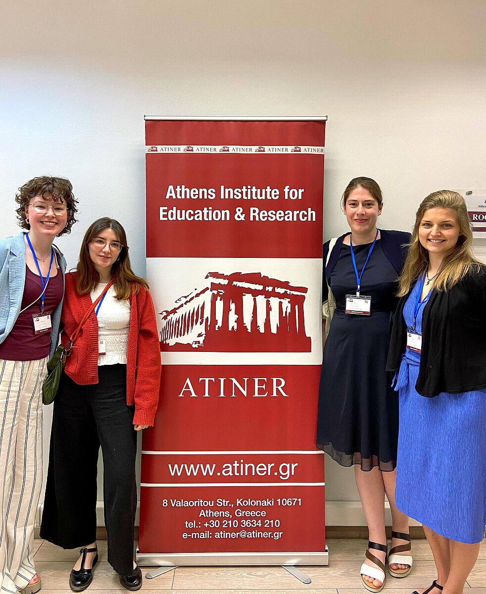 Vier junge Frauen lächeln in die Kamera. Zwischen ihnen befindet sich ein rotes Rollup mit der Aufschrift „Athens Institute for Education & Research“. Die vier Frauen tragen Ausweise an blauen Bändern um den Hals. Zwei von ihnen stehen rechts neben dem Aufsteller, die anderen beiden Personen stehen rechts neben dem Aufsteller.