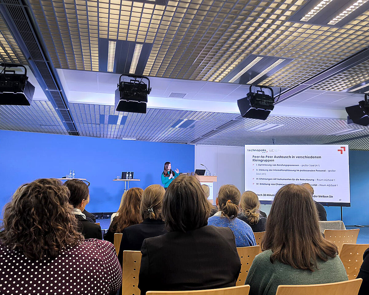 Zu sehen ist ein großer blauer Konferenzraum. Vor einem sitzenden Publikum steht eine Frau an einer Präsentation mit dem Titel "Peer-to-peer Austausch in verschiedenen Kleingruppen".