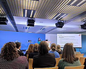 Zu sehen ist ein großer blauer Konferenzraum. Vor einem sitzenden Publikum steht eine Frau an einer Präsentation mit dem Titel "Peer-to-peer Austausch in verschiedenen Kleingruppen".