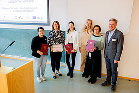 Sechs Personen stehen in einem Lehrraum, hinter Ihnen befinden sich eine Tafel sowie ein großer Monitor. Die Personen halten, gut sichtbar, rote Klappordner in den Händen, auf denen das mecklenburgische Landeswappen in Gold eingeprägt ist.