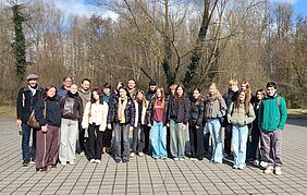 Eine Gruppe junger Menschen posiert für ein Gruppenfoto. Die Personen lächeln freundlich in die Kamera. Sie stehen auf einem gepflasterten Platz, im Hintergrund sind Bäume und strahlend blauer Himmel zu erkennen. Die Personen tragen Jacken, teilweise Schals, Taschen und Rucksäcke.