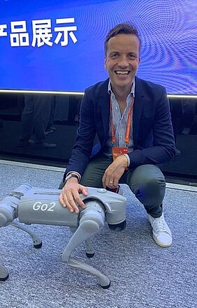 Professor Stefan Schmidt lächelt in die Kamera. Hitner ihm befindet sich eine blaue LED-Wand mit chinesischen Schriftzeichen, vor ihm befindet sich ein hundeartiger Roboter.