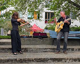 Zwei Personen stehen im Innenhof und musizieren. Die Person im linken Bildrand spielt Violine. Ihre braunen langen Haare wehen ihr ins Gesicht. Die zweite Person steht rechts von ihr. Der Mann spielt Akustikgitarre und sieht freundlich in die Richtung der Violinistin.