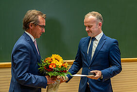 Zwei Männer stehen vor einer grünen Kreidetafel. Beide tragen blaue Anzüge und weiße Hemden. Die linke Person übergibt der rechten Personen einen gelb-orangenen Blumenstrauß, eine Urkunde und eine kleine dunkle Box.