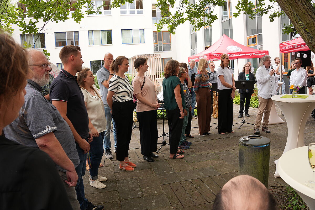 Viele Personen stehen aufgereit nebeneinander. Die Personen unterscheiden sich in Kleidungsstil, Größe, Alter und Geschlecht. Einer von ihnen hält ein Mikrofon und spricht zu einem Publikum.