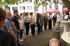 Viele Personen stehen aufgereit nebeneinander. Die Personen unterscheiden sich in Kleidungsstil, Größe, Alter und Geschlecht. Einer von ihnen hält ein Mikrofon und spricht zu einem Publikum.
