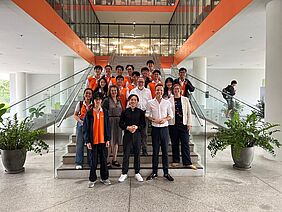 Zu sehen ist ein Gruppenfoto in einem großen, hellen Foyer. Die anwesenden Personen verteilen sich in vier Reihen auf einer großen Treppe. Die jungen Personen befinden sich vorrangig in den hinteren Reihen und tragen orangefarbene Shirts. Im Hintergrund befinden sich architektonische Elemente wie Säulen, Gelände, Fenster und Türen aus Glas und Stein. Die Szenerie ist vorrangig in weiß und orange gehalten. 