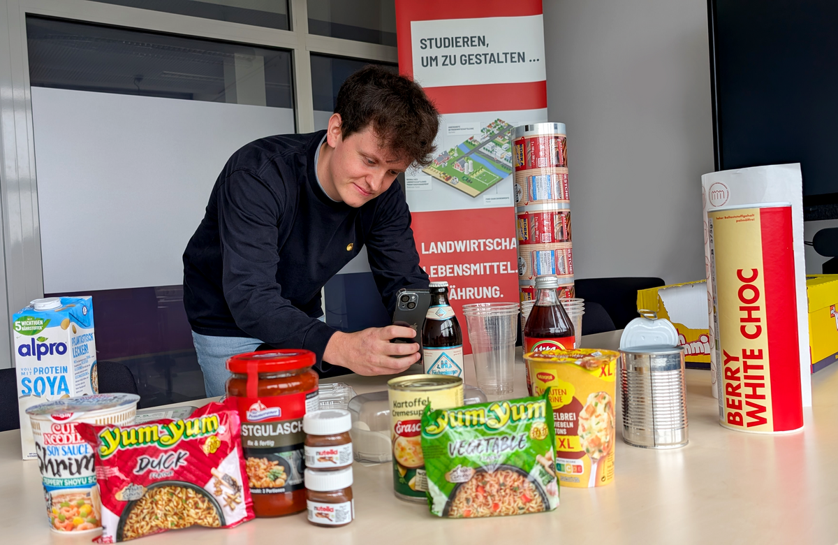 Ein junger Mann mit welligem dunklem Haar und dunklem Langarmpullover scannt mit seiner Handykamera verschiedene Produktverpackungen von Getränken und Instant-Produkten wie Ramen oder Kartoffelsuppe. Im Hintergrund befindet sich ein Aufsteller mit der Aufschrift „Studieren, um zu gestalten“.