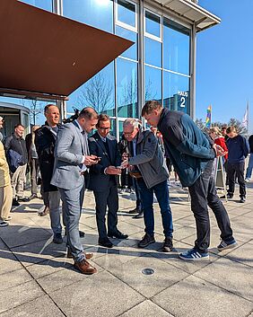 Drei Männer schauen, im Halbkreis stehend, auf ihre Smartphones. Hinter ihnen steht eine weitere Person mit Kameraequipment, neben ihnen schaut ein Mann nach vorne gebeugt ebenfalls auf die Handydisplays. Am Boden vor den Personen befindet sich der eingelassene Navigationskontrollpunkt.