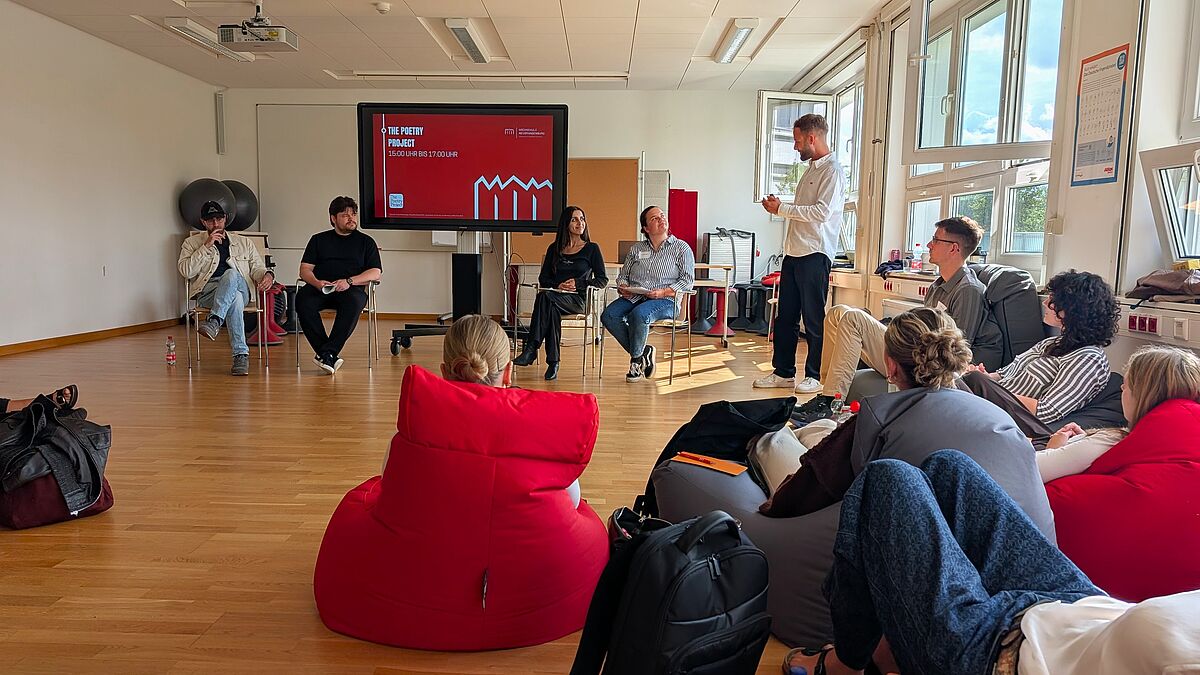 Vier Personen sitzen vor einem Monitor auf Stühlen. Neben ihnen, an einer Fensterfront, steht eine weitere Person, die zu ihnen spricht. Auf dem Monitor ist die Aufschrift "The Poetry Project" zu erkennen. Zu den Personen zugewandt sitzen weitere Menschen, wahrscheinlich eine Klasse, auf verschiedenfarbigen Sitzsäcken.