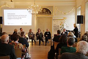 Ein festlicher, freundlich-heller Raum. Im Raum befinden sich Stühle, auf denen Personen sitzen, die nach vorne in Richtung einer Podiumsdiskussion schauen. Die Diskussionsteilnehmenden sitzen ebenfalls auf Stühlen, mit Blick zum Publikum. Hinter den Expert*innen befindet sich eine Leinwand mit einer Präsentation, die auf die Diskussionsrunde aufmerksam macht. Außerdem befindet sich ein länglicher Aufsteller neben den Diskussionsplätzen, auf dem „Tag der Kulturfördervereine“ steht.