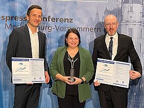 Professor Dr. Schmidt, Ministerin Stefanie Drese und Minister Christian Pegel posieren gemeinsam vor einem blauen Aufsteller der das Schweriner Schloss und die Aufschrift "Landespressekonferenz Mecklenburg-Vorpommern" zeigt.