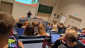 Gestikulierend spricht eine Person zu einem gut gefüllten Hörsaal. Das Publikum hat sich auf blauen Hörsaalstühlen auf verschiedenen Etagen verteilt und schaut nach vorne. Einige Per-sonen haben Laptops vor sich aufgeklappt.