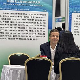 Professor Schmidt sitzt einer vermutlich weiblichen, langhaarigen Person gegenüber. Hinter ihm befindet sich ein Aufsteller mit chinesischen Schriftzeichen und dem Aufdruck "Hochschule Neubrandenburg - University of Applied Sciences".