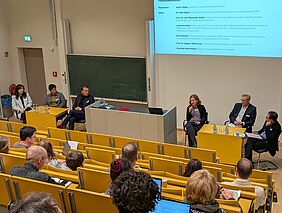 Sechs Personen sitzen an zwei Tischen, vorne in einem Hörsaal. Eine Frau, die sich unter den Podiumsteilnehmenden befindet, redet in ein Handmikrofon. Hinter ihr sind die Namen der vorne sitzenden Personen auf weißem Hintergrund zu sehen. Die Personen sitzen dem Hörsaalpublikum zugewandt.