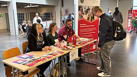 An einem Tisch sitzen drei Personen. Vor ihnen schaut eine weitere Person interessiert auf den Tisch, auf dem sich Flyer befinden. An dem Tisch klebt ein DIN-A4 großer Zettel mit der Aufschrift "Fachschaft SBE".