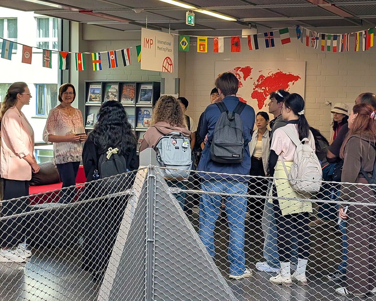 Ein Zusammentreffen verschiedener Personen – junge Menschen mit Rucksäcken schauen auf zwei förmlich gekleidete Personen, die zu ihnen sprechen. Im Hintergrund befindet ein rotes Sofa, ein offenes Bücherregal und eine Leinwand mit einer Weltkarte.