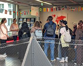 Ein Zusammentreffen verschiedener Personen – junge Menschen mit Rucksäcken schauen auf zwei förmlich gekleidete Personen, die zu ihnen sprechen. Im Hintergrund befindet ein rotes Sofa, ein offenes Bücherregal und eine Leinwand mit einer Weltkarte.