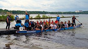 Links und rechts von einem Steg legen zwei Drachenboote an. Beide Boote sind gefüllt mit Menschen. Sie halten Paddel in das wellige Wasser. Auf dem Steg stehen mehrere blau gekleidete Personen vom Sportclub Neubrandenburg.