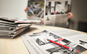 Auf einem Tisch liegt ein Stapel Zeitungen, davor eine aufgeklappte Ausgabe mit dem Titel „Forschung und Transfer“ und einem roten Kugelschreiber auf der Titelseite. Unscharf im Hintergrund ist eine Person zu erkennen, die die Zeitung, wohl das Forschungsjournal der Hochschule Neubrandenburg, liest.