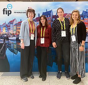 Vier junge Frauen lächeln freundlich in die Kamera. Hinter ihnen befindet sich ein Aufsteller, der auf den FIP Weltkongress für Pharmazie und Pharmazeutische Wissenschaften in Kopenhagen hinweist.