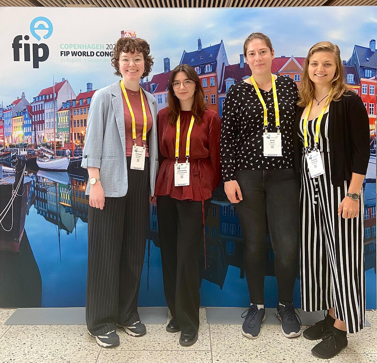 Vier junge Frauen lächeln freundlich in die Kamera. Hinter ihnen befindet sich ein Aufsteller, der auf den FIP Weltkongress für Pharmazie und Pharmazeutische Wissenschaften in Kopenhagen hinweist.