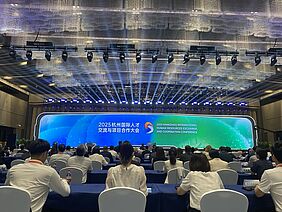 Zu sehen ist eine große LED-Wand mit der Aufschrift "Hangzhou International Human Resources Exchange and Cooperation Conference" sowie viele sitzende Menschen, die in Richtung LED-Wand schauen.