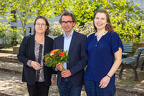 Die Vorsitzende des Senats Claudia Vogel und die Stellvertretende Vorsitzende Susanne Dreas gratulieren Gerd Teschke zur Wiederwahl und überreichen ihm einen bunten Blumenstrauß auf dem sonnigen Innenhof des Hochschulhaupthauses.